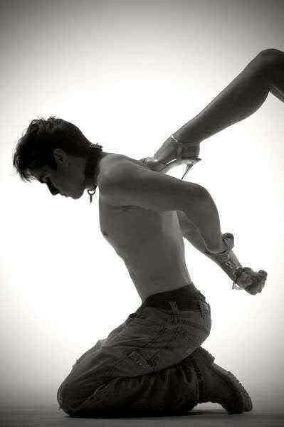 Man in jeans and a collar kneeling. A woman's leg is lifted, stiletto-heeled foot pressing between his shoulder blades.