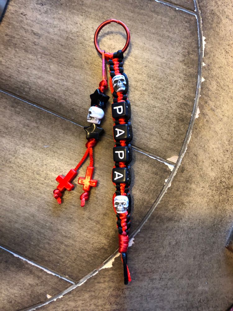 A picture of a bead and macrame keychain sitting on a wooden table. The keychain has two parts. It’s made on a red metallic keyring. The first is a longer, square knotted keychain made with red and black satin thread. The word Papa is spelt out with four beads. There are two white skull beads; one above and one below the word. It is tied off with a red gathering knot. The second is a smaller knotted keychain attached with a keychain hook. It’s made with red thread and starts with a knot, two black star beads holding a white skull bead, knotted again, and finished off with two strands. The strands are individually ended with red upside down cross beads finished with a decorative knot. 