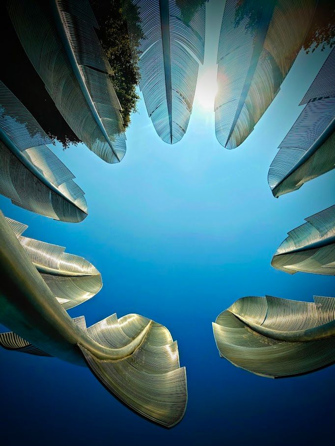 The photo captures a striking upward view of the Choctaw Native American monument *Kindred Spirit*, featuring a ring of towering, stylized steel feathers reaching toward a vivid blue sky. The feathers, rendered with fine, parallel lines, gleam in the sunlight, creating a shimmering, almost ethereal effect. The perspective from below frames the open sky like a starburst, with sunlight peeking through the top, evoking a sense of reverence and connection to the heavens. The monument’s design blends grace and strength, symbolizing resilience, unity, and the enduring spirit of the Choctaw people. 