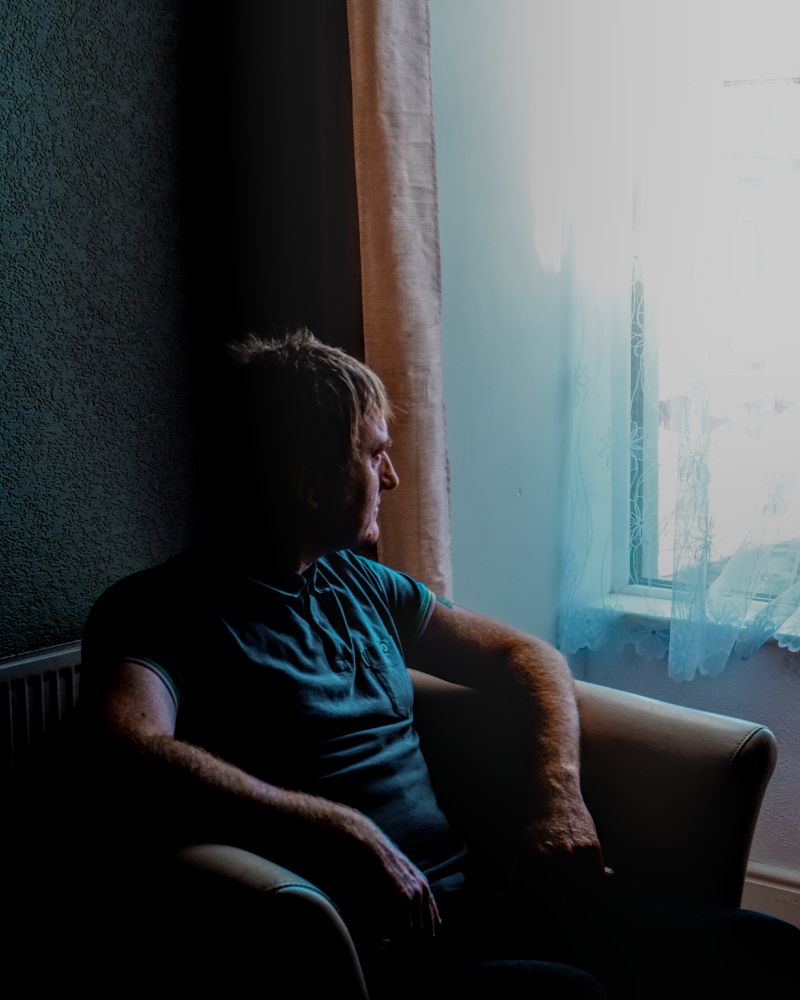 This photograph captures a man seated in an armchair beside a window, gazing contemplatively toward the light streaming in. The right side of his body and face are softly illuminated by the daylight, while the rest of his form recedes into deep shadow, creating a striking contrast between brightness and darkness. He wears a teal polo shirt, and his relaxed posture, with one arm resting on the chair and the other across his lap, conveys a sense of quiet introspection or waiting. The curtain and lace by the window diffuse the light, giving the scene a gentle, almost painterly atmosphere that emphasizes mood over detail.