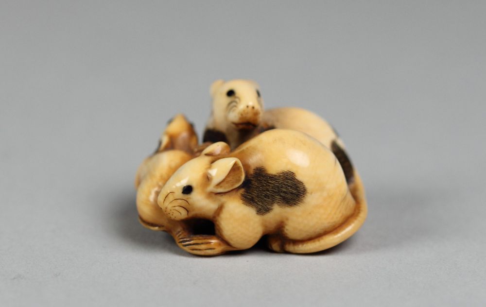 An ivory netsuke of three rats. One rat faces to the left, one faces away from the viewer, and one peeks over the back of the one in front. They have black eyes and dark, fur-textured patches. They all have details such as ears, whiskers, tails, paws, and noses. The piece is about an inch tall. 