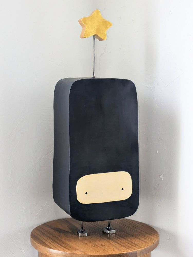 a  black rectangular figure with a little mask and a chunky gold star sprouting from its head