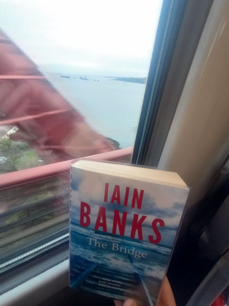 A copy of Iain Banks' novel The Bridge, photographed from a train carriage travelling over the Forth Rail Bridge, with a large, blurry red girder visible in the background. 
