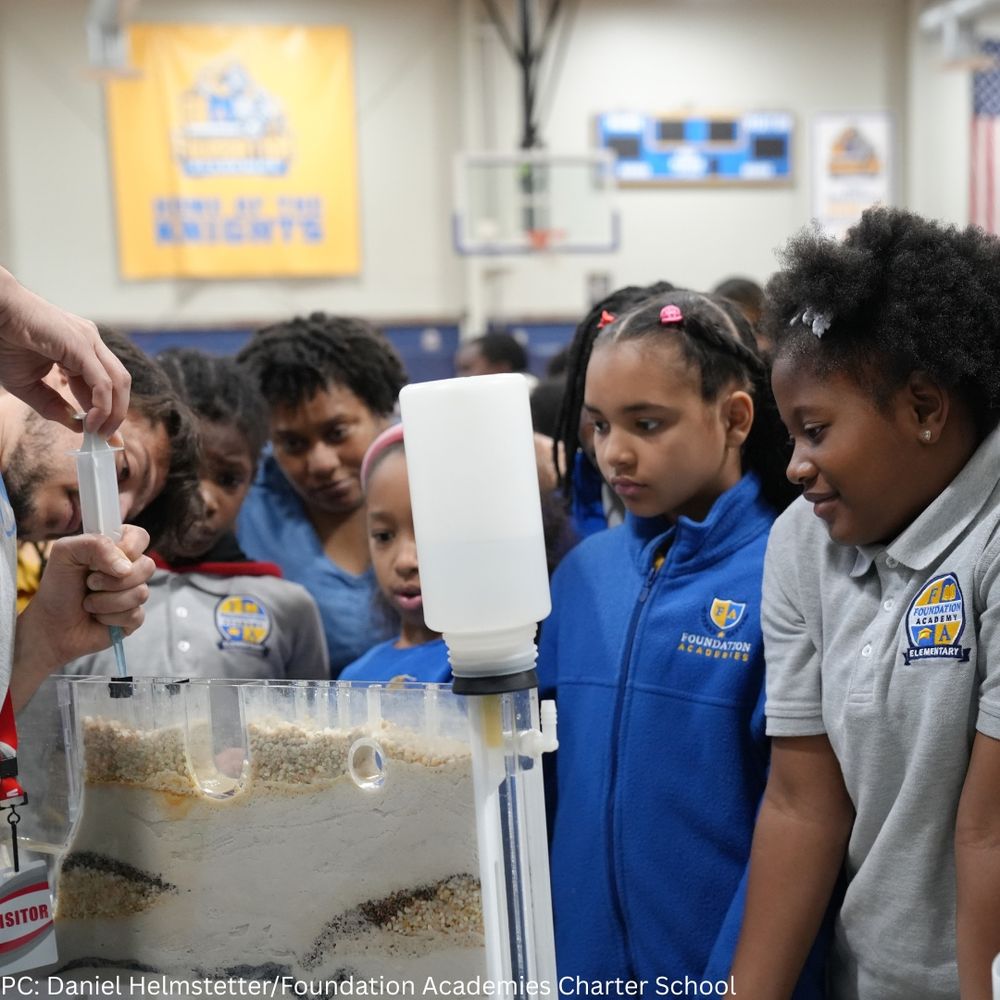 A group of students from Foundation Academies Charter School gather around an interactive groundwater model demonstration. The model shows layers of sand and soil, and an adult uses a syringe to “pump” liquid from the model, simulating water movement. The students watch attentively, engaged and curious. 