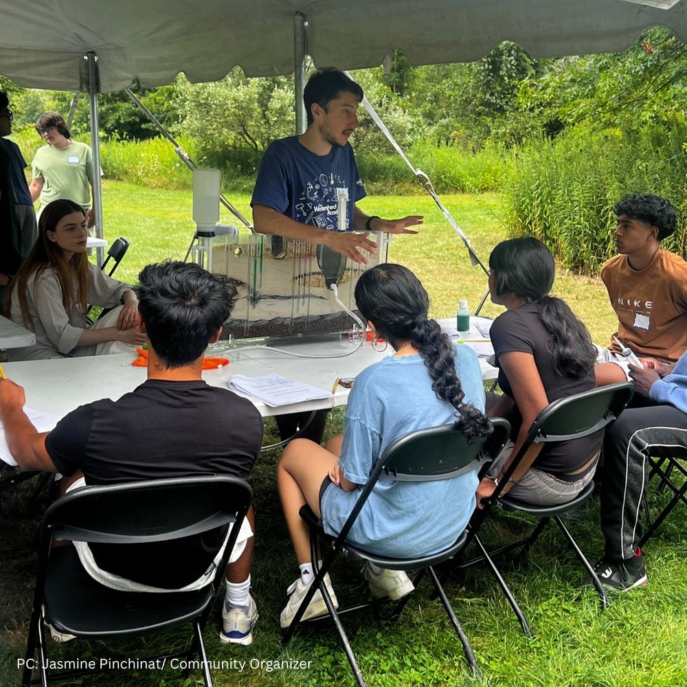 A young man in a blue t-shirt stands under a tent, gesturing as he explains a groundwater model to a group of students seated around a table. The model, a clear box filled with layers of soil and water, sits in front of him. The students, holding papers and pens, listen attentively. The outdoor setting is lush with greenery in the background.