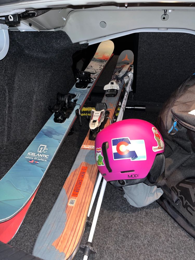 Photo of two pairs of skis, a set of poles, and a helmet with a merged trans&colorado flag sticker (and a sticker of Kermit with a lesbian flag) in the back of a car 