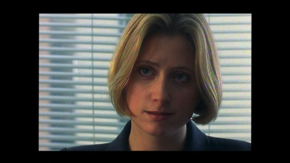 A screenshot from the 2001 DVD. A close-up of a blonde, white woman sitting in front of some office blinds. There are numerous odd rainbow colour artefacts showing in the details of her hair.