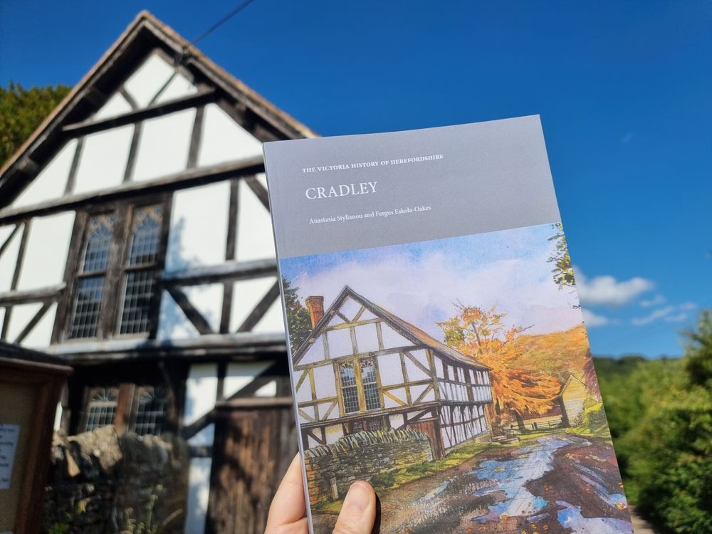 A book, 'Cradley', by Anastaisia Stylianou and Fergus Eskola-Oakes, with a cover featuring a timber framed building in an autumnal landscape. The gable end of the same building, Cradley parish hall, formerly the church ale hall, built in the 15th century is the backdrop, below a deep blue sky.