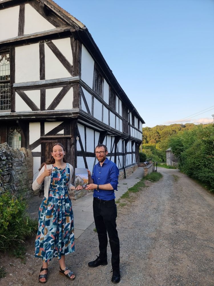 The authors, Anastasia Stylianou (left), in a long dress, smiling, and Fergus Eskola-Oakes, wearing a blue shirt and black jeans, holding their book against the backdrop of Cradley parish hall, a timber framed building which is also on the cover.