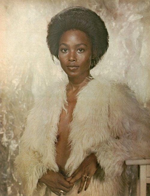70s Actress Brenda Sykes in a long, furry white coat and nothing else. She looks calmly bemused. 