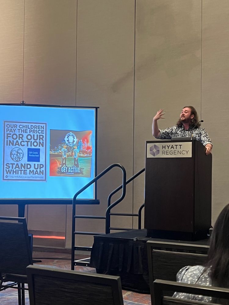 Reed Van Schenck, comm scholar, wearing a patterned shirt, in front of a lecturn, gesturing to a PowerPoint slide featuring white nationalist content found on Telegram
