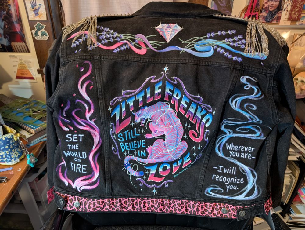 Photo of the finished paint job on the back of my Freaks jacket. Top panel: a diamond, stalks of lavender, and a ribbon; center panel:  "Little Freaks Still Believe in Love" written in a  shield shape around a pink tiger wrapped in strings of pearls and surrounded by sparkles; Left panel: pink and purple fire around the lyric "Set the world on fire" from Strange; right panel: blue and white smoke around the lyric "Wherever you are, I will recognize you," from Lavender Love.  it's painted in Pink, Blue and purple and trimmed in hot pink leopard print ribbon with metal spiked epaulettes.