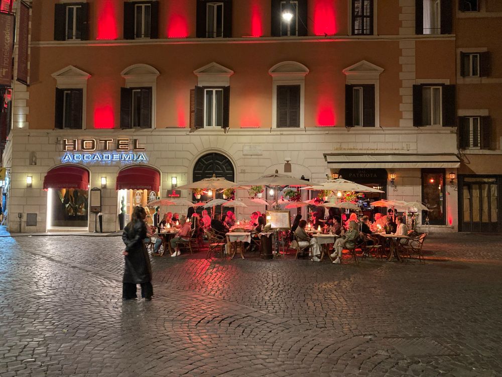 In a small piazza in front of Hotel Accademia. The building has red uplights between the windows on the second and third floors. In the piazza there is restaurant seatjng with some red lights on the guests. A restaurant host, all in black in the open space of the piazza, invites passersby to dine. 