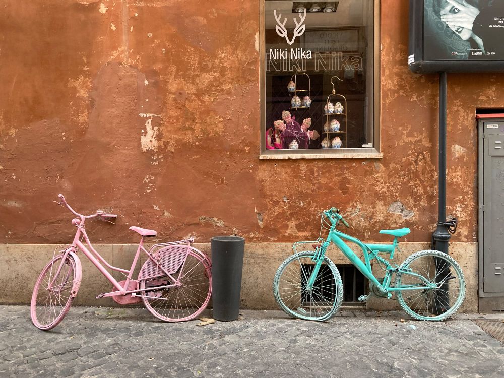 In an alleyway, right of center is a shop window. In the bottom half of the photo there are two broken bikes leaning against the wall- on painted all pink and the other painted all teal. 