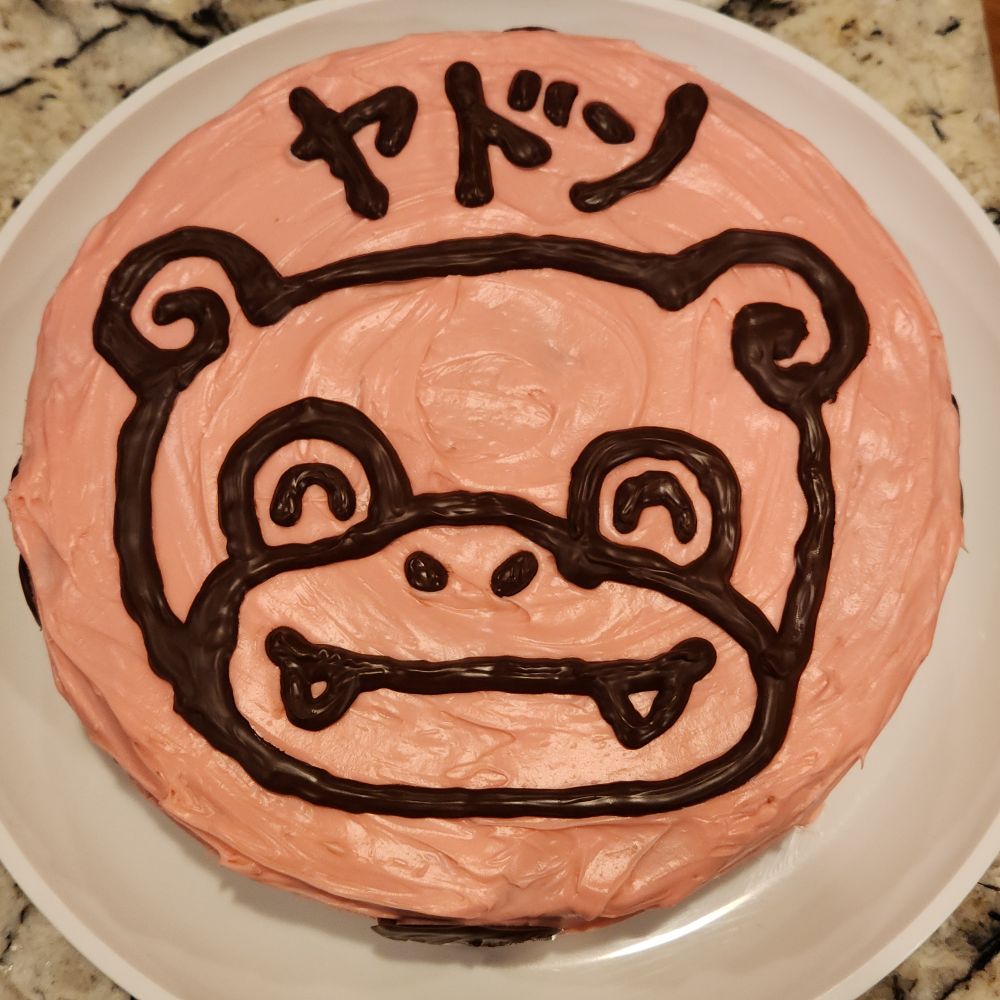 A round cake topped with pink frosting and chocolate pieces of a Slowpoke's face and its Japanese name 