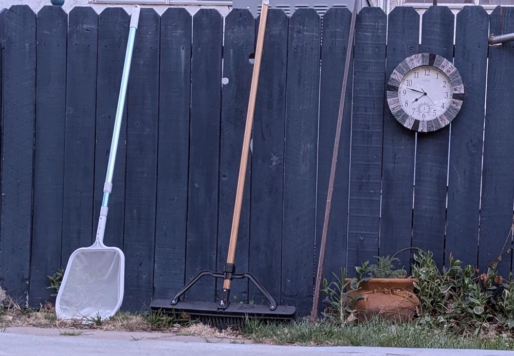 A clock and three gardening tools against a deep navy fence.