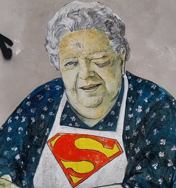 Bild/ Foto zeigt Wandbild von Seniorin mit kurzen grauen Haaren, sitzend mit weißer Schürze mit Supermann-Emblem darauf, lächelnd und mit dem linken Auge zwinkernd.