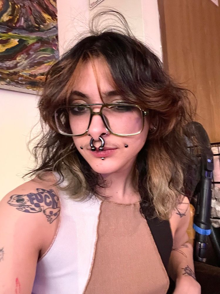 the body of the dogpile system, a white person with black wavy hair, tattoos, facial piercings and glasses. it has thick black eye makeup around its round brown eyes, and is winking. the lighting in the room is very pink