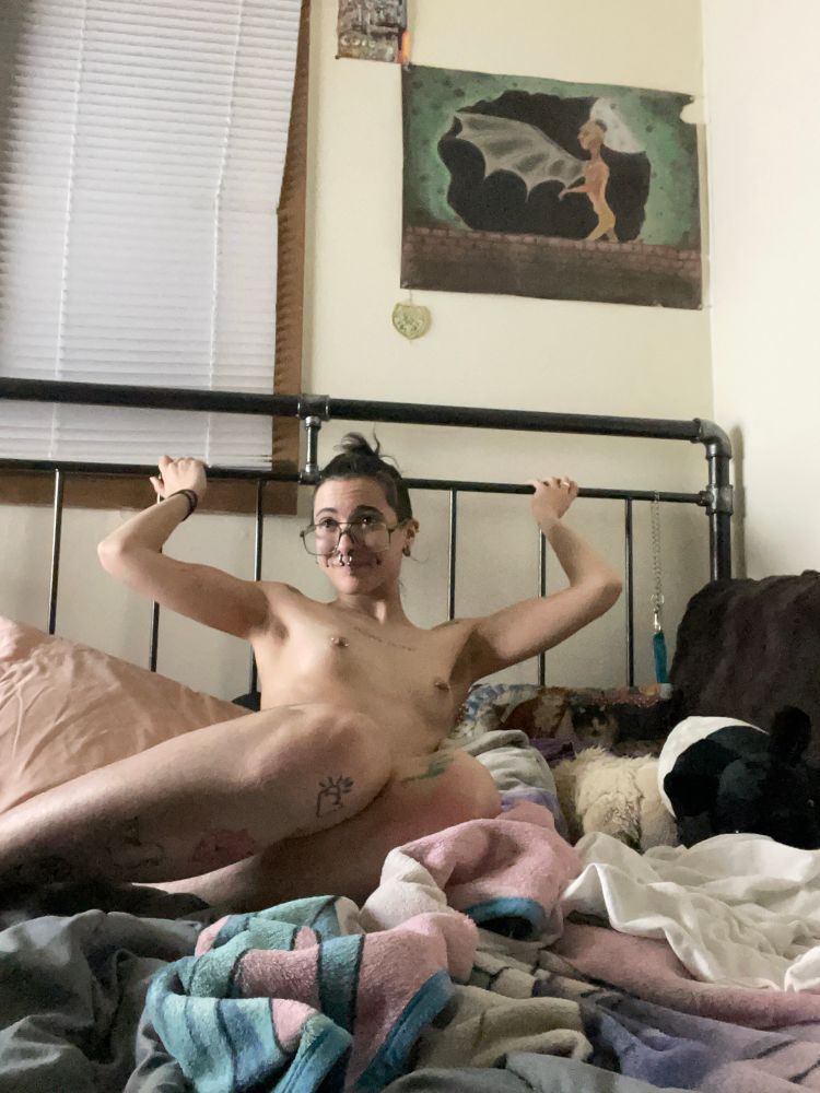 the body of dogpile, a white person with visible tattoos, and face and body piercings, is sitting in a comfy pile of blankets in his bed. his bedframe is made of metal, and both hands reach above his shoulders to grab it. 
it is looking up nervously, but with its legs pulled to cover its naked body (lower than his tits), like a cornered animal during play time hehehehe. 