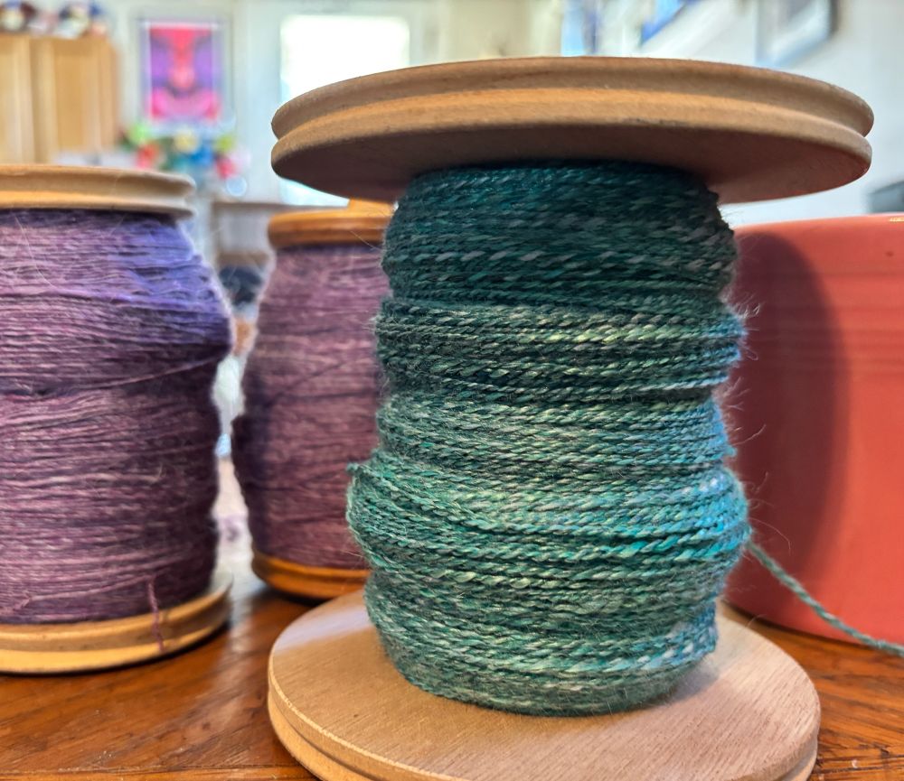 3 bobbins of fiber. 1 with a green blend that is plied and 2 with a purple blend (singles, waiting to be plied.) 