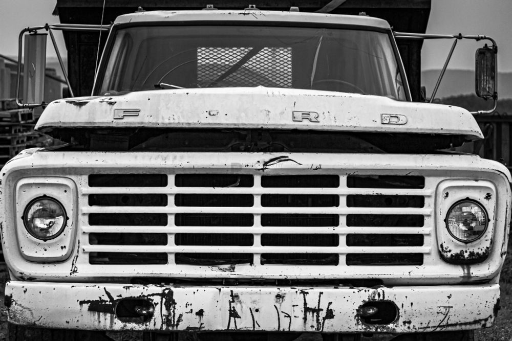 Monochromatic front shot photo of a old Ford pickup truck, with a visible aftermarket dump contained attached to it. The hood of the pickup is popped open, and there are scratches, dents, and rust all over it.