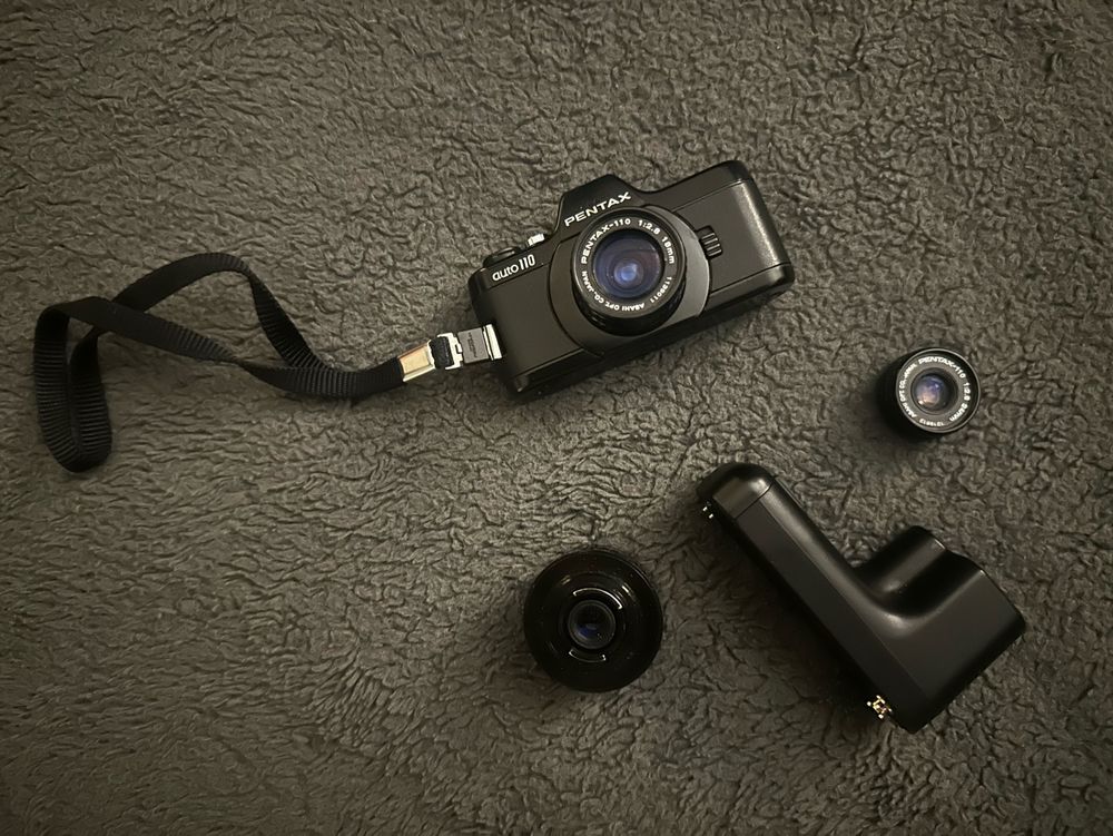 Pentax Auto 110 film camera, lying on its back, with two lenses and the auto film winder on a flat surface for display. 