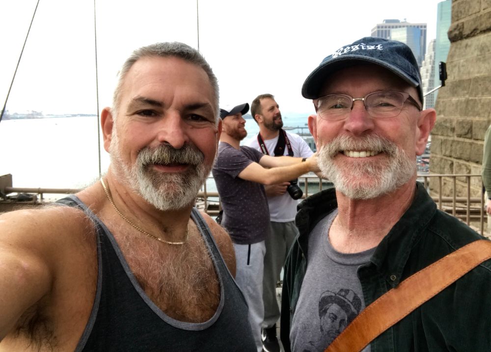 As originally captioned when I first posted it seven or eight years ago, we saw this handsome bearded couple taking a selfie on the Brooklyn Bridge and decided to do the same. My husband and I, both bearded, one wearing a ball cap, pose on the bridge — behind us, two men, both bearded, one wearing a ball cap, pose for the phone camera in the outstretched arms of one of them. 