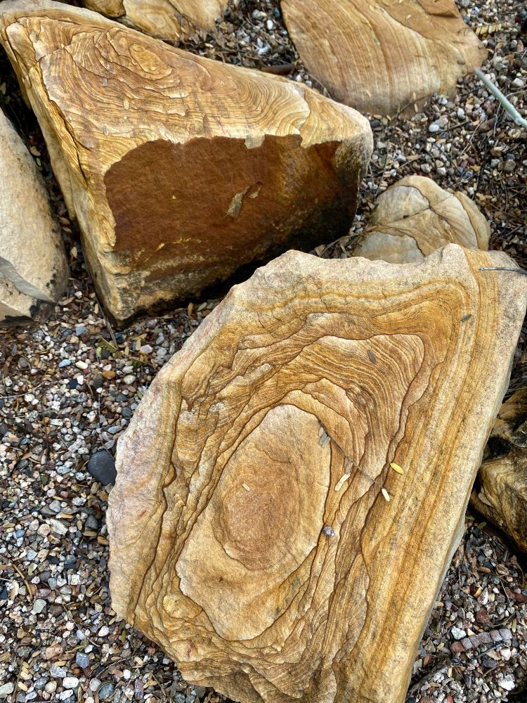 Cool broken sedimentary rocks with twisting strata in ochres, tans, and browns on a bed of water-tumbled gravel. 