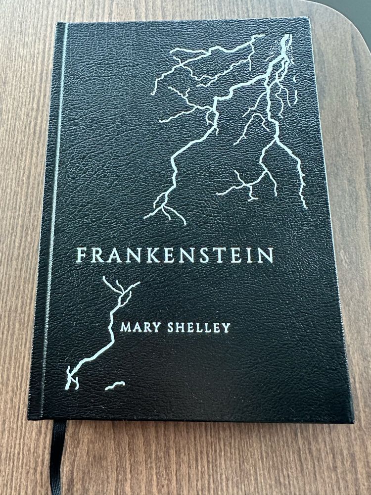 Photo of a book on a table: Mary Shelley’s Frankenstein. The hardback cover is glossy black and the title is written at the bottom in sharp white text, while jagged lines of white go from the upper right down to the bottom left of the book like forks of lightening