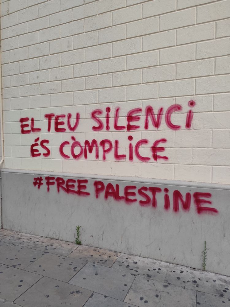 Pintada a una paret on hi diu en lletres vermelles "el teu silenci és còmplice #freepalestine"