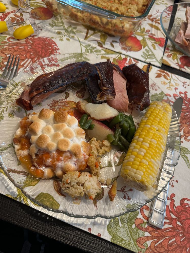 Photo of a dinner plate full of food, on a floral printed placemat. Food pictured: Turkey wing, corn on the cob, stuffing, green beans, diced potato and sweet potato casserole which is orange potato mash with roasted marshmallows on top. 