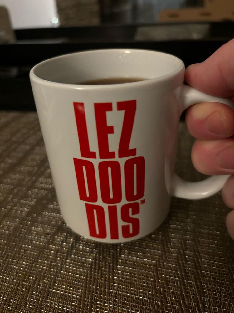 A white ceramic coffee mug, full of steaming coffee. On the side of the mug are large red letters that boldly proclaim LEZ DOO DIS 