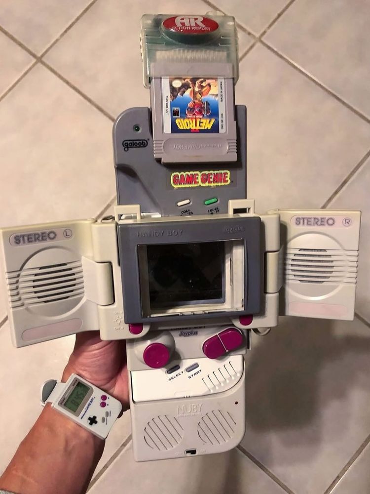 A person is holding an original Nintendo Game Boy that has been heavily modified with various attachments. The Game Boy has a large gray accessory attached, labeled “HANDY BOY,” which adds extra stereo speakers (labeled “STEREO L” and “STEREO R”) that fold out from the sides, as well as a magnifying lens over the screen. Below the screen are large, red joystick-style buttons labeled “Joyplus.” Attached to the game slot is a gray Game Genie cheat device with a sticker that reads “GAME GENIE” in red and yellow. A Metroid II: Return of Samus game cartridge is inserted into the Game Genie, and on top of that is an Action Replay device. The person is wearing a wristwatch designed to look like a miniature Game Boy, with a small screen and buttons. The background shows light-colored tiled flooring.