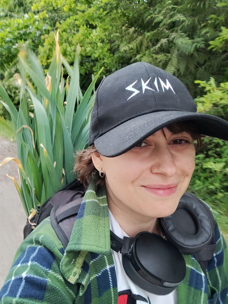 A white woman with brown hair and eyes wearing a black hat that says "Skim" and big black headphones around her neck. She is carrying a bunch of green garlic in a backpack and it sticks out behind her head. 