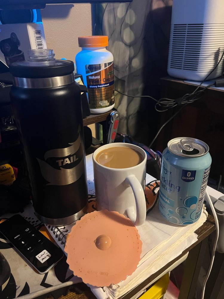 In the foreground, A black water bottle, a full cup of coffee with creamer and an open can of sparkling water on my desk. In the background there is a bottle of fiber capsules.