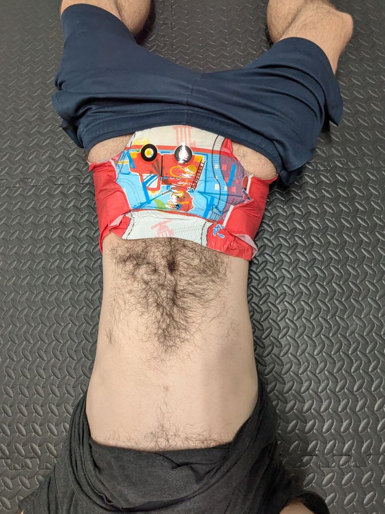 Fuzzy man laying on a gym floor, shirt pulled up to show his belly and shorts pulled partway down to show his soggy fire truck adult diaper