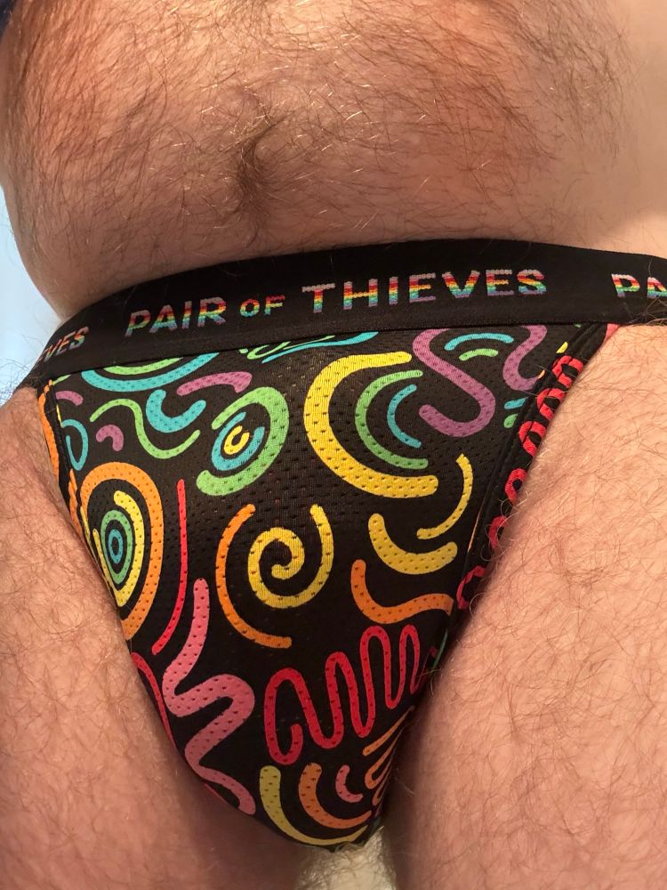 A white, hairy man with a small belly wearing a Pair of Theives Pride-themed jockstrap. The waistband is black, with Pride striped lettering. The jock is black with Pride rainbow squiggles and semi-circles