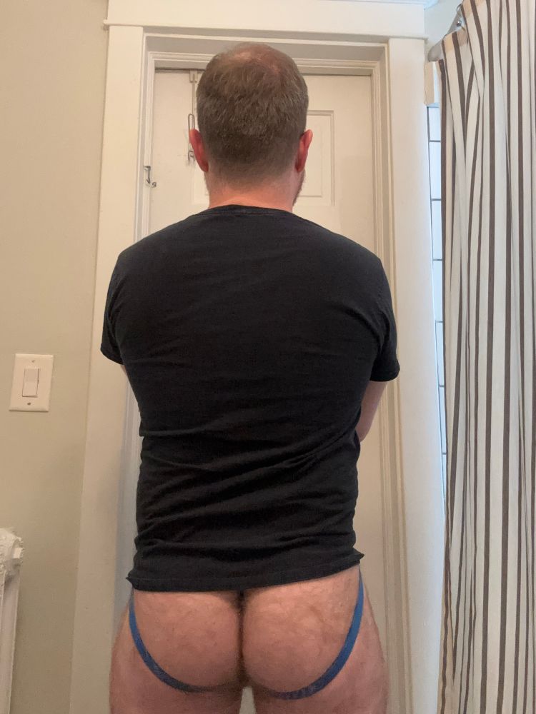 A white, middle-aged, cisgender man with brown hair, with his back to the camera. He is wearing a black shirt and a blue jockstrap. He has a hairy ass. The color scheme of the bathroom he is standing in is white.