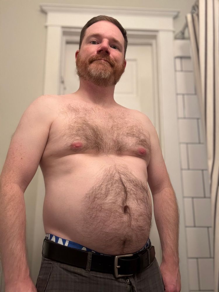 A ginger-gray-bearded, brown-haired middle-aged white man standing shirtless. He has body hair on his stomach, near his pecs, and on his arms. He is wearing brown shorts and a belt. The blue waistband of his Andrew Christian underwear is showing 