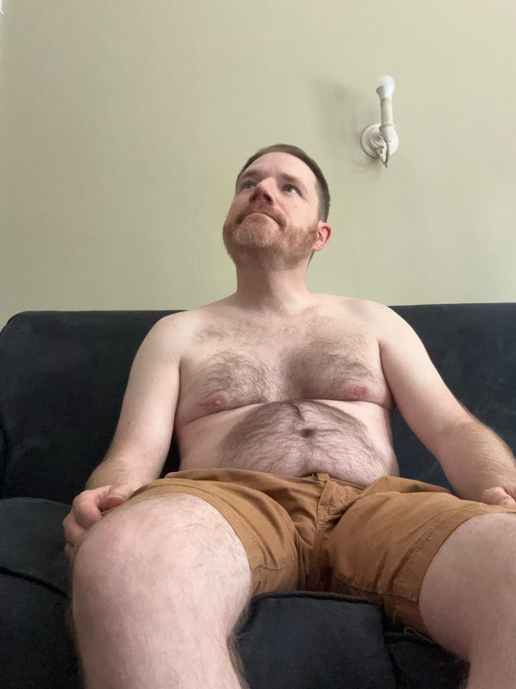 A white man with brown hair and a ginger gray beard sitting shirtless on a navy blue couch. He is wearing almond colored shorts. His legs are also hairy. He has hair around his pectorals and his belly. There is an antique light on the white wall.
