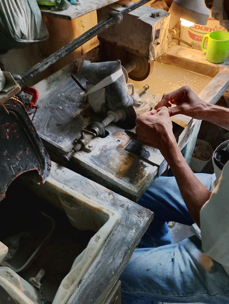 Close nas mãos do pedrista manuseando algo pequeno numa serra diamantada de disco num estúdio caracteristicamente sujo