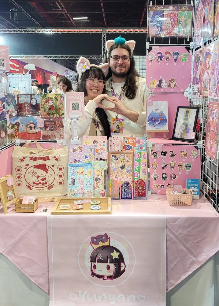 Mesa de evento, exposição de produtos com pronta, ecobag, adesivos, bottons. Presença da artista e seu marido fazendo um coração com as mãos