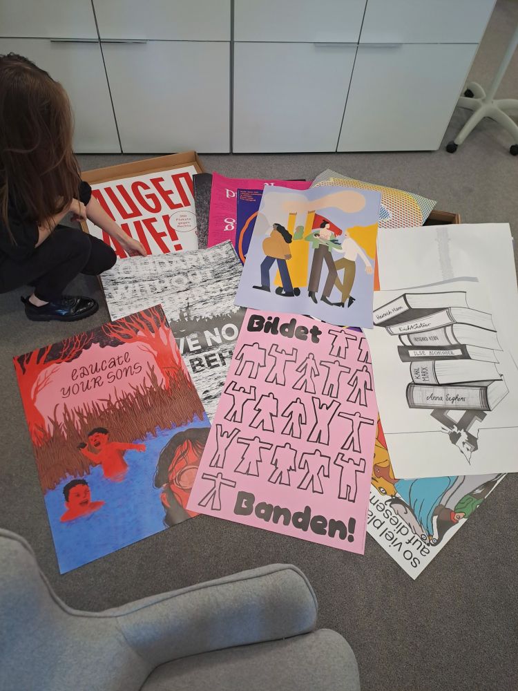 Eine Person hockt auf dem Boden und betrachtet mehrere bunte Plakate, die auf dem Teppich ausgebreitet sind. Darauf sind unterschiedliche politische und künstlerische Motive und Slogans zu sehen, unter anderem „Educate your sons“, „Bildet Banden!“ und "Solidarity without borders - Leave no one behind" sowie Illustrationen von Menschen und Büchern.