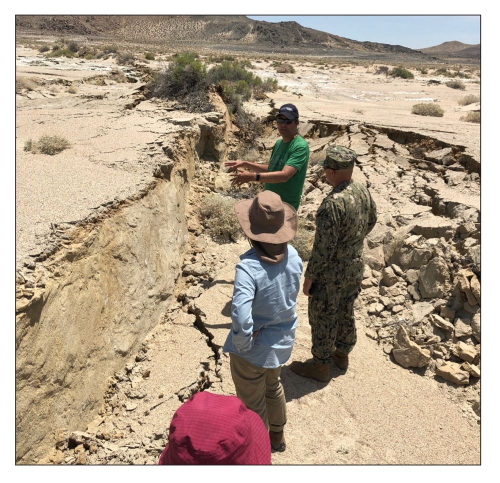 3 people stand in the desert looking at a small cliff (scarp) formed by the earthquake. The ground around them is cracked and broken. 