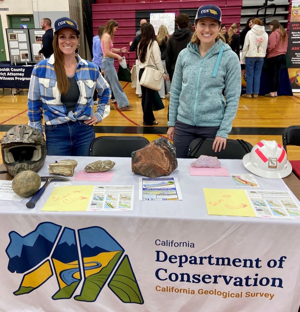 Two smiling women wearing Cgs baseball hats stand behind a table. On the table or rocks, a rock hammer, a hardhat, a helmet and various informational pamphlets. The table runner has the logo of a bear and says California Department of conservation California geological survey.