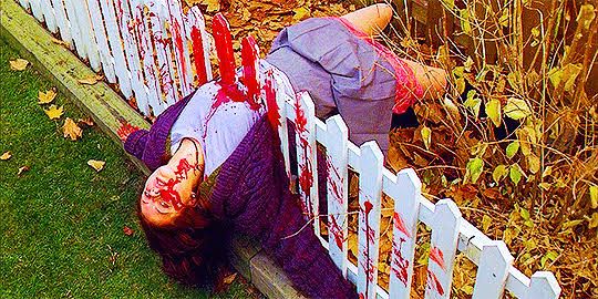 Cena de filme Possuída (ginger snaps) em que menina branca adolescente usando uniforme de escola está com o corpo desmaiado atravessado por uma cerca, coberta de sangue (mesmo dentro do filme, a cena é falsa, algo que ela fez pra parecer um cenário de terror)