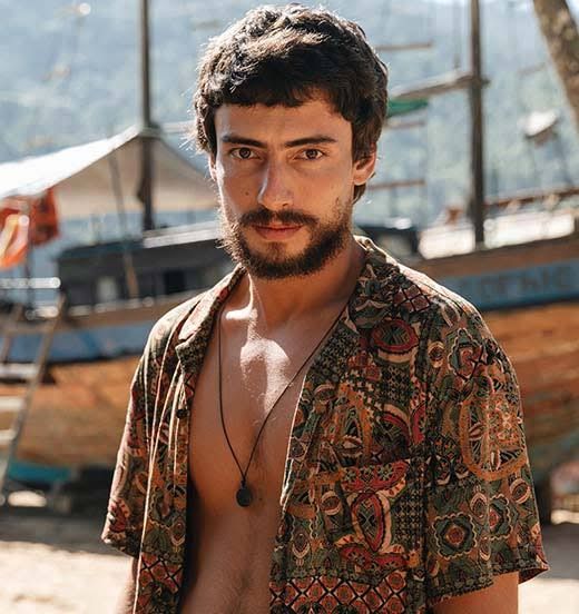 Mesmo homem descrito ao lado, dessa vez com um blusão colorido e estampado aberto sem nada por baixo (peito, barriga e braços a mostra), um cordão que vai até o centro do peito, usando barba e bigode e com o cabelo cortado curto (sem óculos)