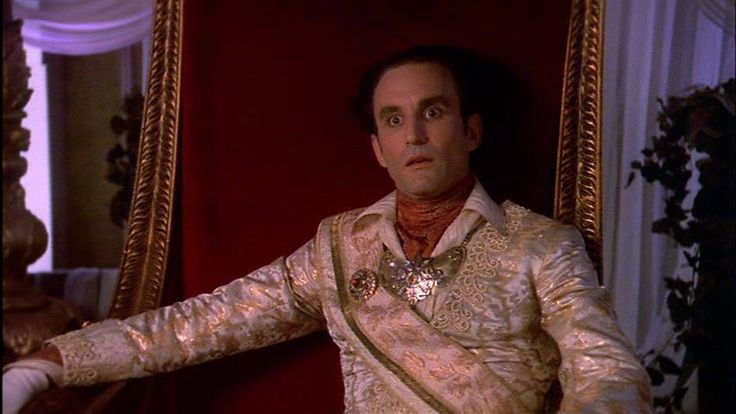 Emperor Cartagia, a man wearing an ostentatious coat in white and gold as well as a large golden medallion, sitting on a golden throne with red upholstery, looking surprised.