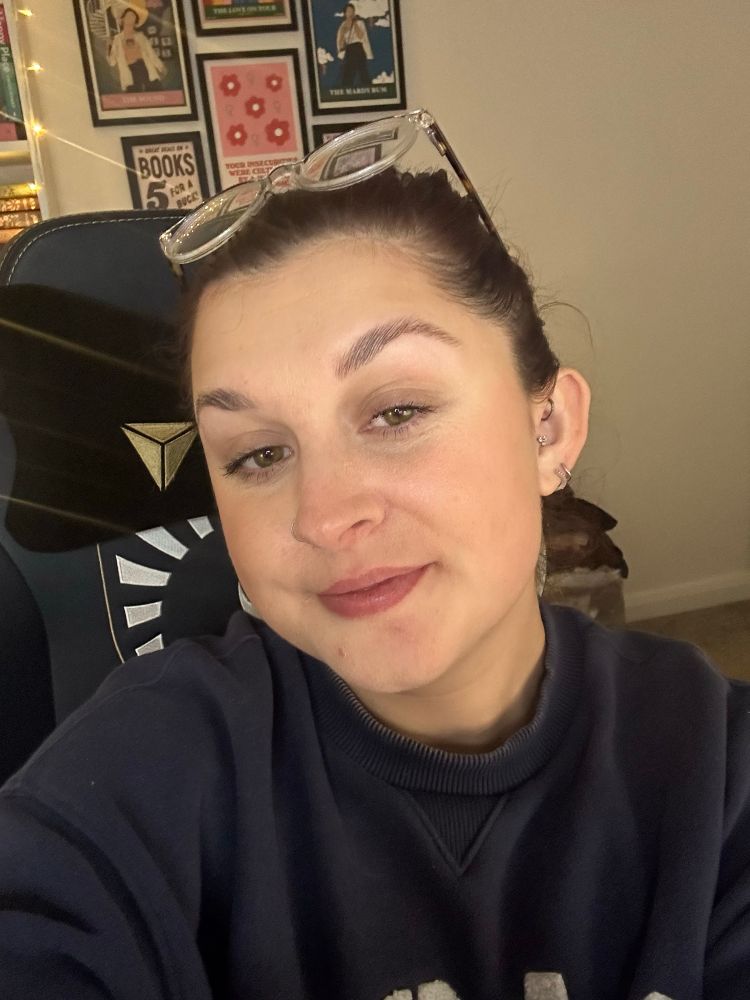 Kira smiling with her mouth closed. Wearing a navy blue jumper and with her glasses on top her head. She has her hair tied back off of her face