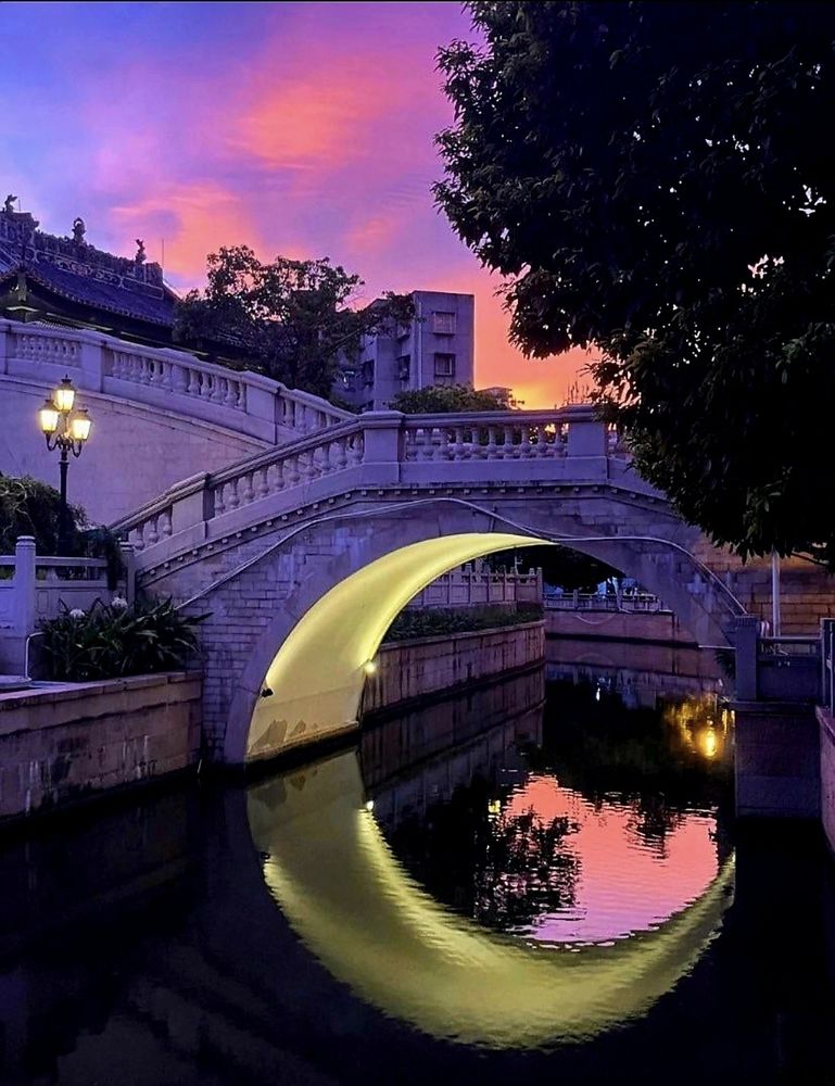 Image of a bridge over a canal with underlights, its reflection  creates the shape of a crescent moon, a purple tone sunset in the sky, solitary and calm feel